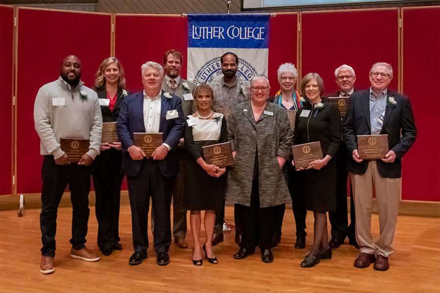 Ten Luther alumni holiding award plaques stand on a stage with Luther president Jenifer K. Ward