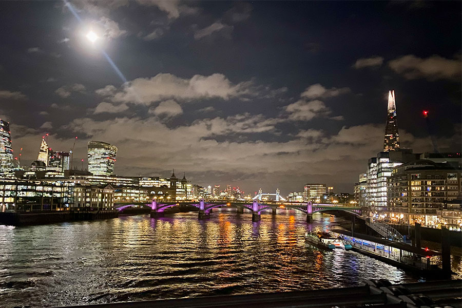 A lit-up nighttime city skyline and bridge over a river, the moon above