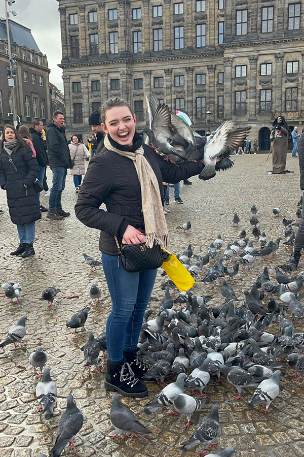 A laughing woman in a plaza with dozens of pigeons at her feet and several pigeons perched on her outstretched arm