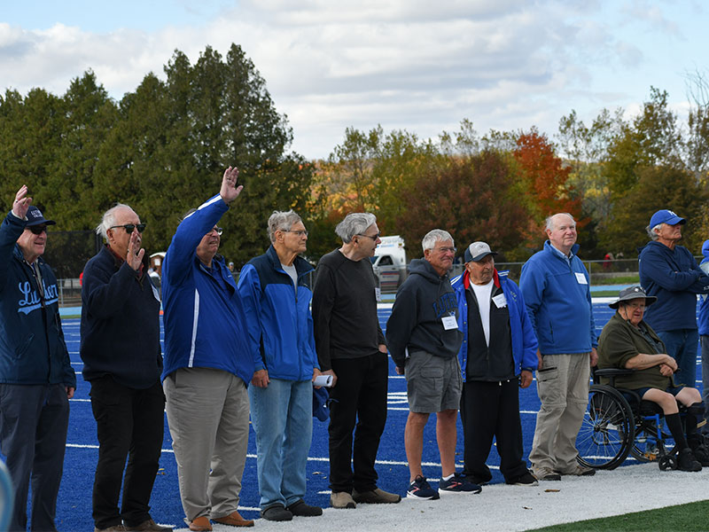 A line of older adult men standing on a football field with blue turf