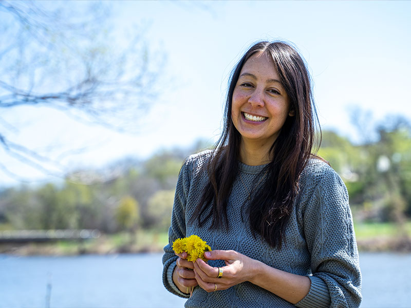 A smiling woman in front of open water holding a bouquet of dandelions