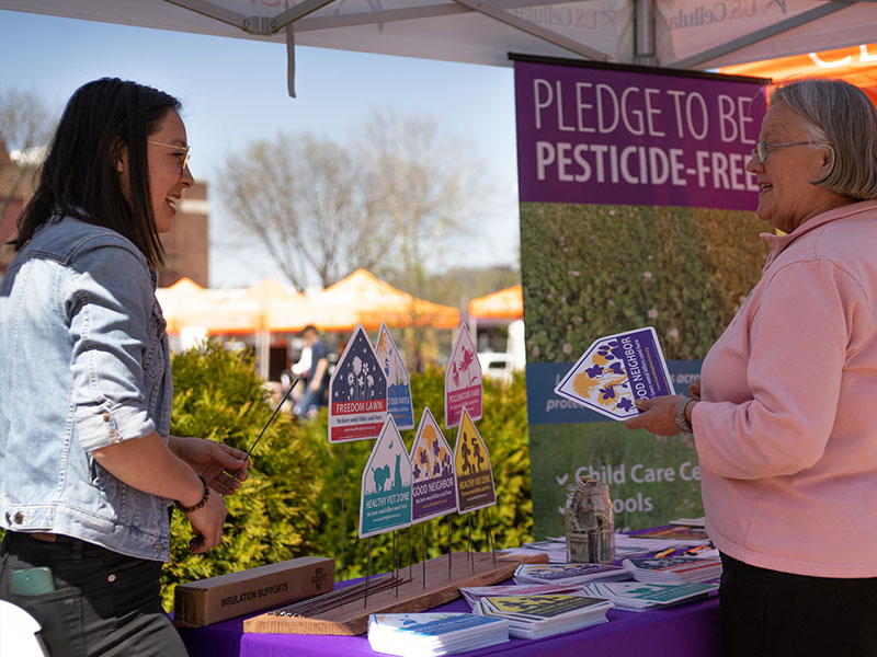 Two women chatting in an outdoor canopied booth that contains a poster and brochures about pesticides