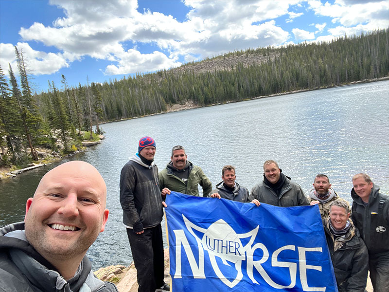 Eight men standing at the edge of a lake holding a Luther Norse banner