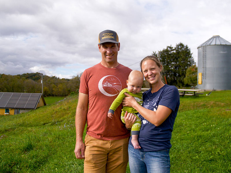 A man, woman, and young child stand outdoors with a silo and low farm building in the background