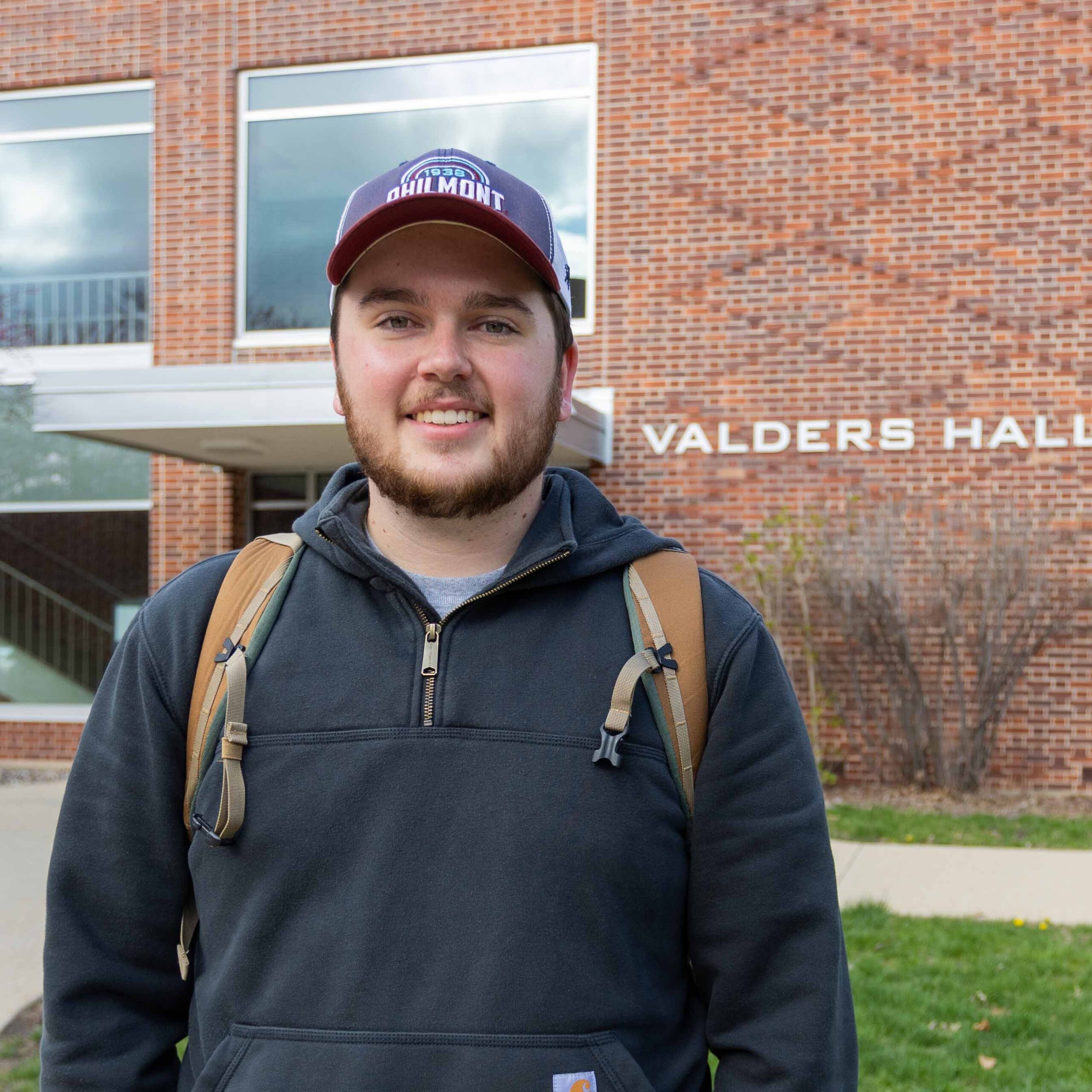 Cole wears a hat and a backpack, standing in front of Valders Hall.