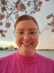 Emily Hursh in glasses and a pink sweater against a sunset on the water. 